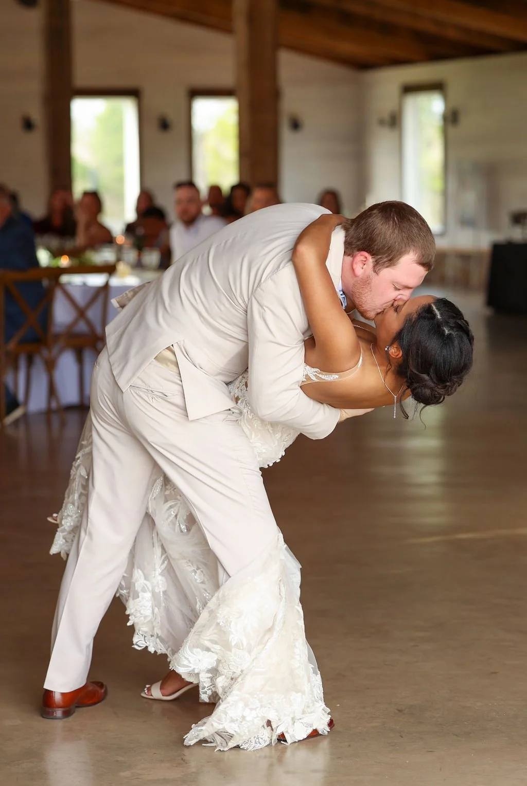 A newlywed couple sharing a kiss during their wedding dance. The groom is in a white suit, and the bride is in a white lace wedding gown, dancing in a rustic reception hall with guests seated at tables in the background.