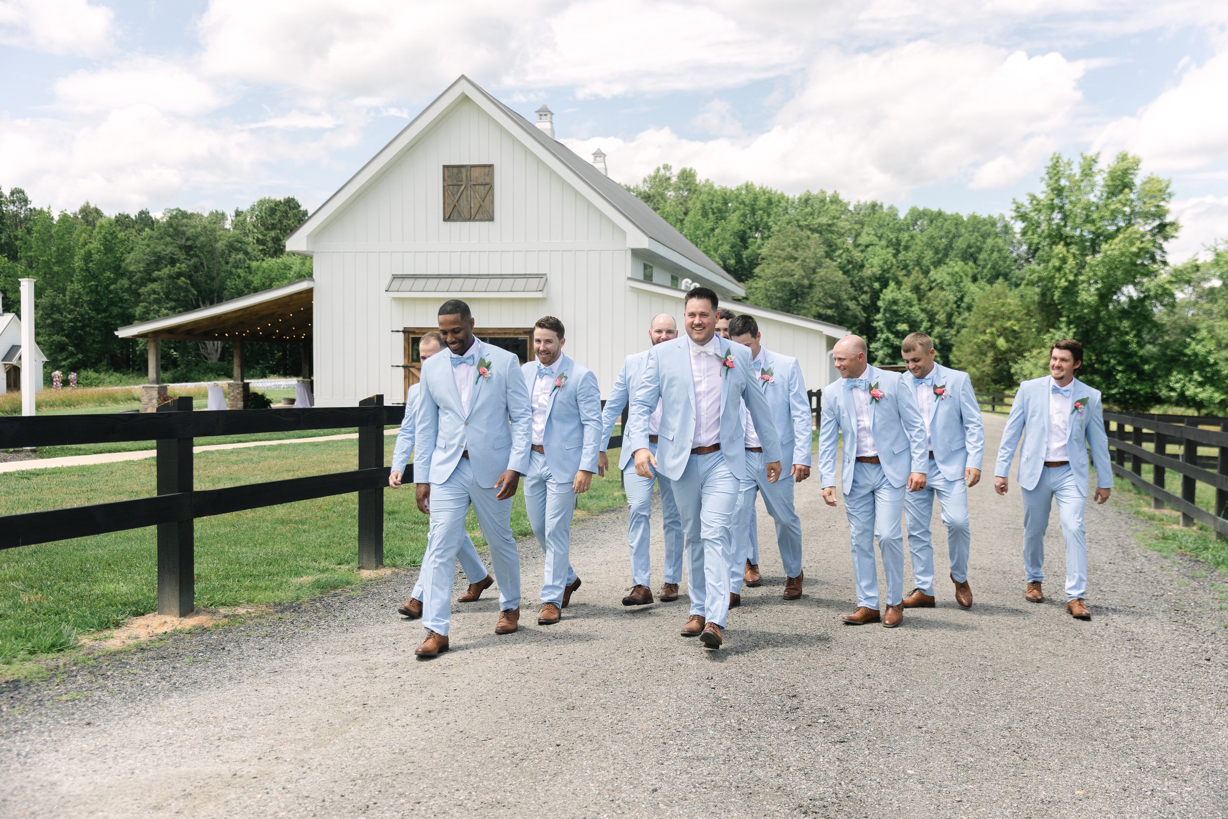 A group of men dressed in light blue suits walking outdoors on a gravel path, with a white barn, grassy field, and trees in the background.