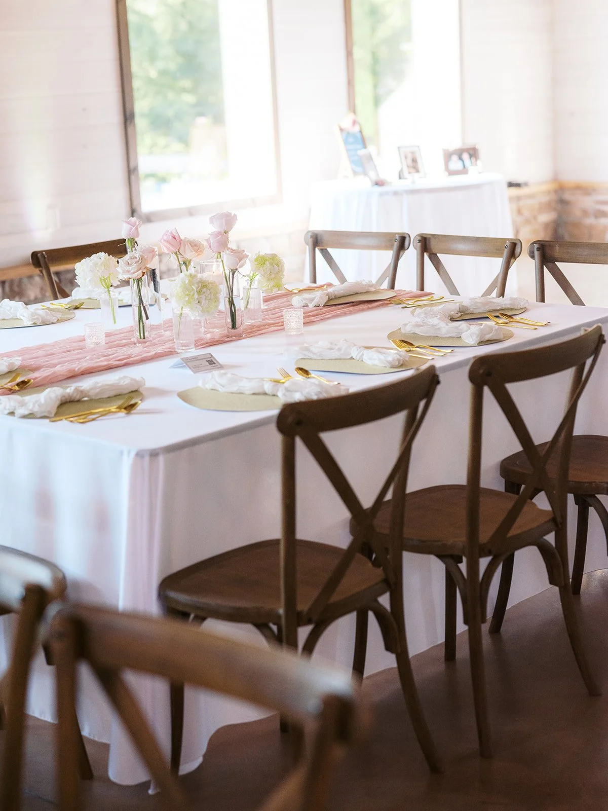 Wedding reception table setup with white tablecloth, pink runner, floral centerpieces, and place settings with gold utensils and white napkins, in a bright room with wooden chairs and large windows.