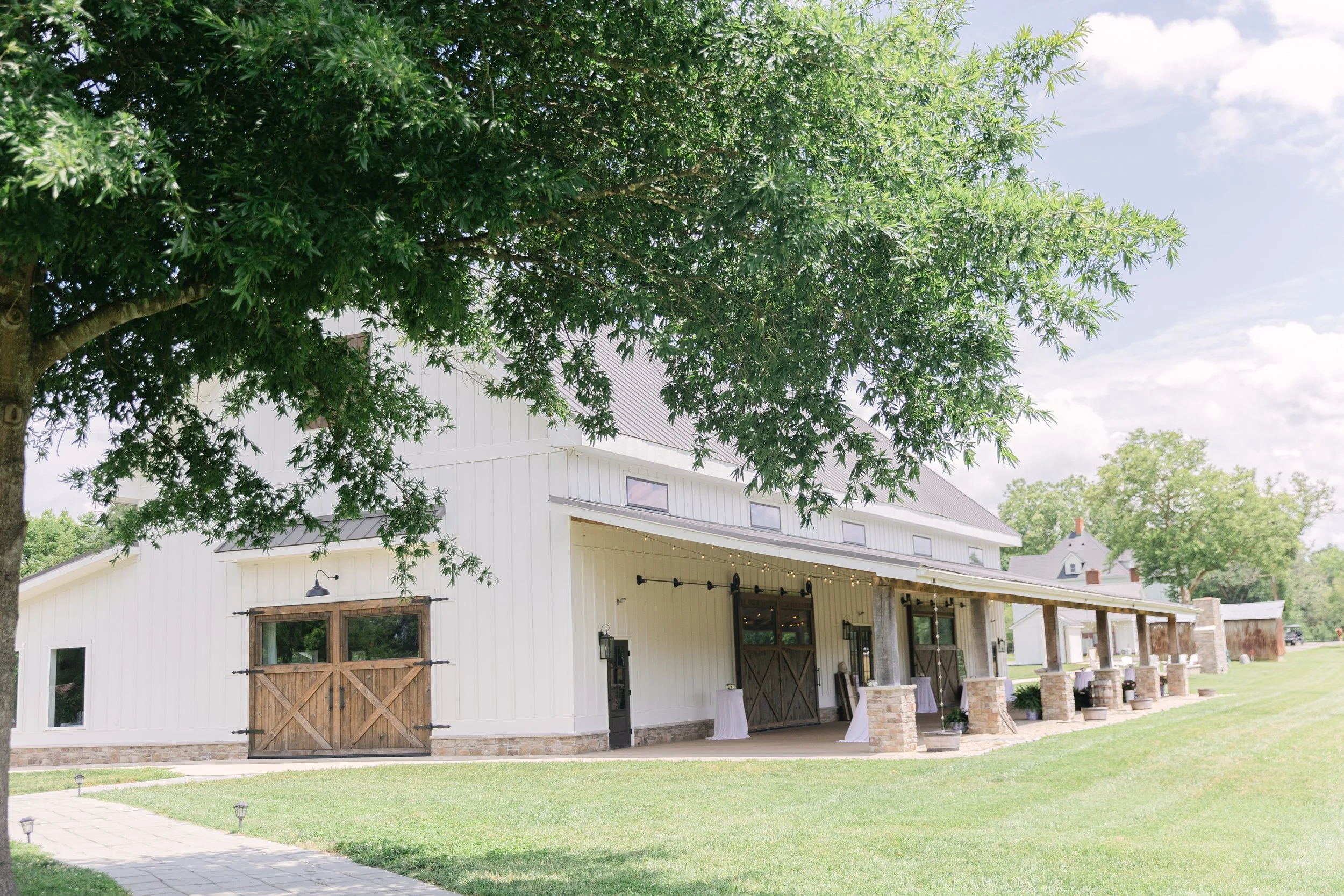 A white barn-style building with wooden accents, surrounded by a green lawn and trees on a sunny day.