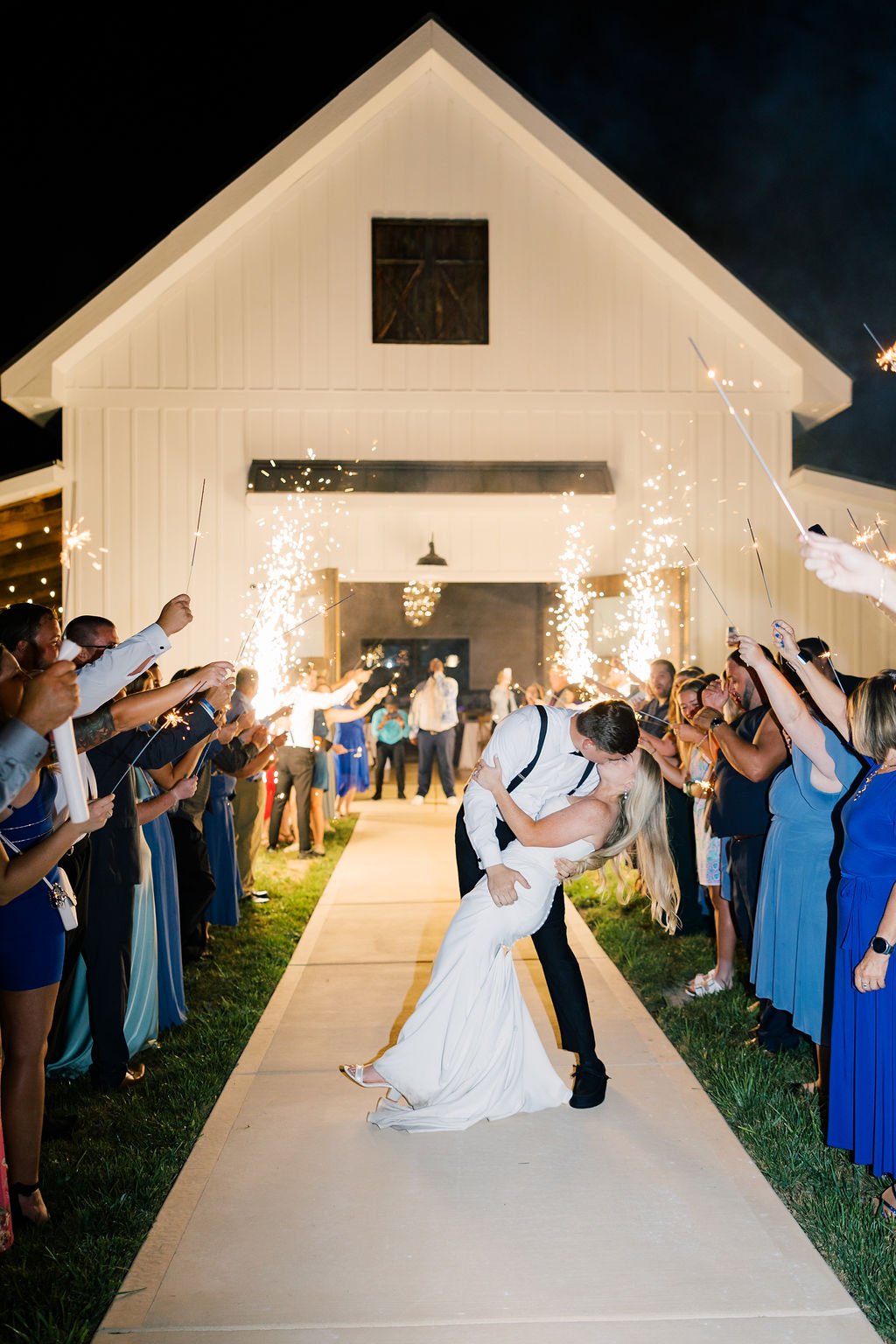 A newlywed couple shares a kiss while being surrounded by friends and family holding sparklers during a night celebration outside a white barn-style building.