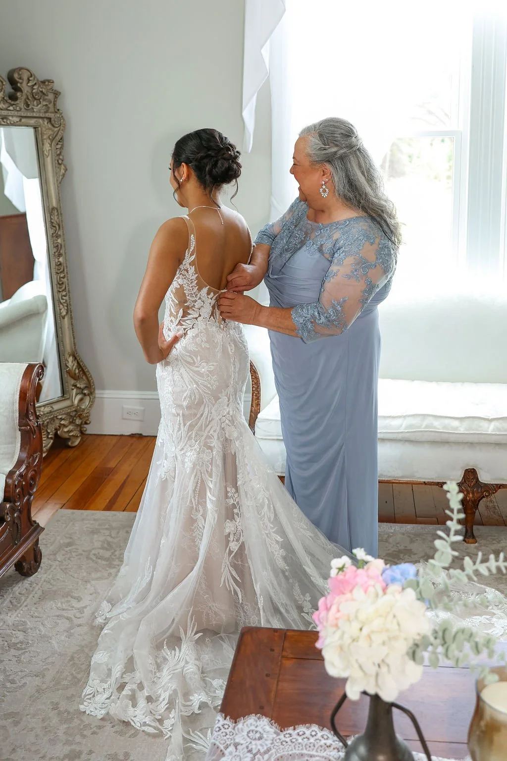 A bride getting dressed in her wedding gown with help from an older woman in a blue dress, in a bright room with window, mirror, and flowers.