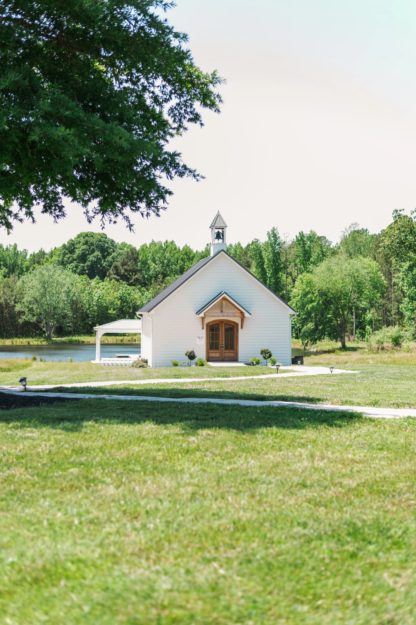 A small white church with a wooden door, surrounded by green grass and trees, near a pond, under a clear sky.