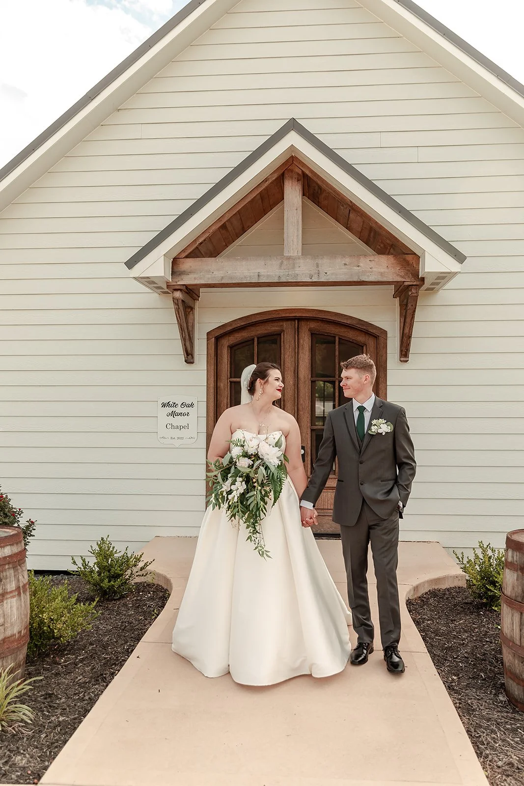 A bride and groom holding hands and looking at each other in front of a small chapel with a wooden door and white siding.