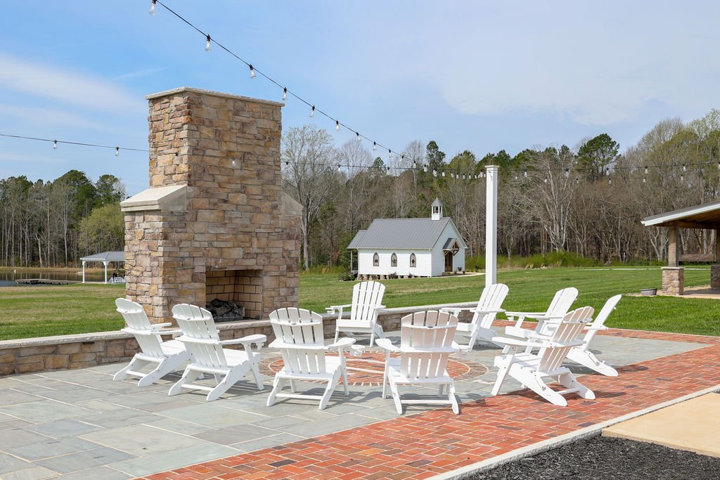 Outdoor patio with white Adirondack chairs arranged in a circle around a stone fireplace, on a paved surface with string lights overhead and a small white chapel in the background.