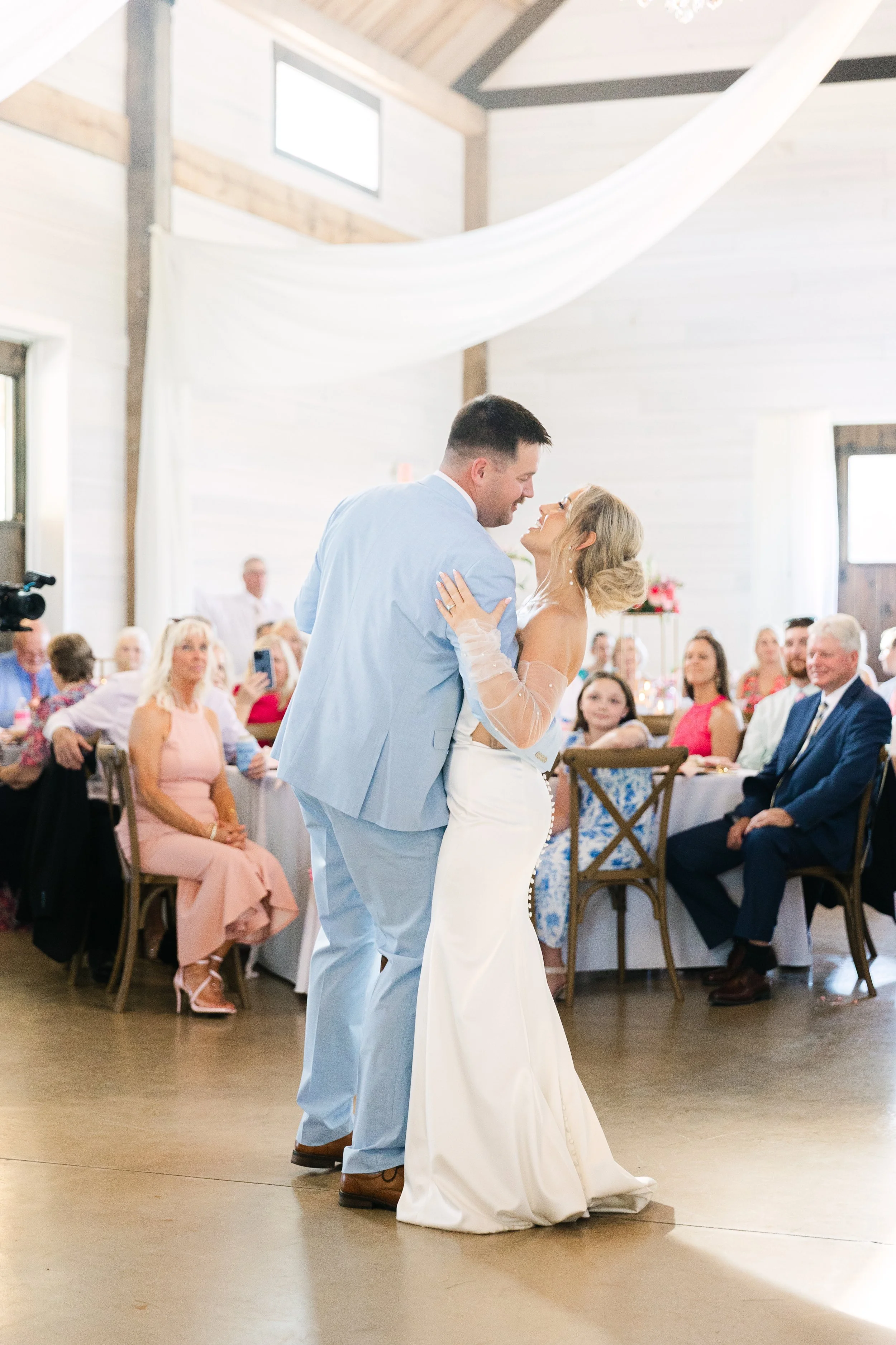 Bride and groom dancing at their wedding reception in a decorated venue with guests seated at tables watching.