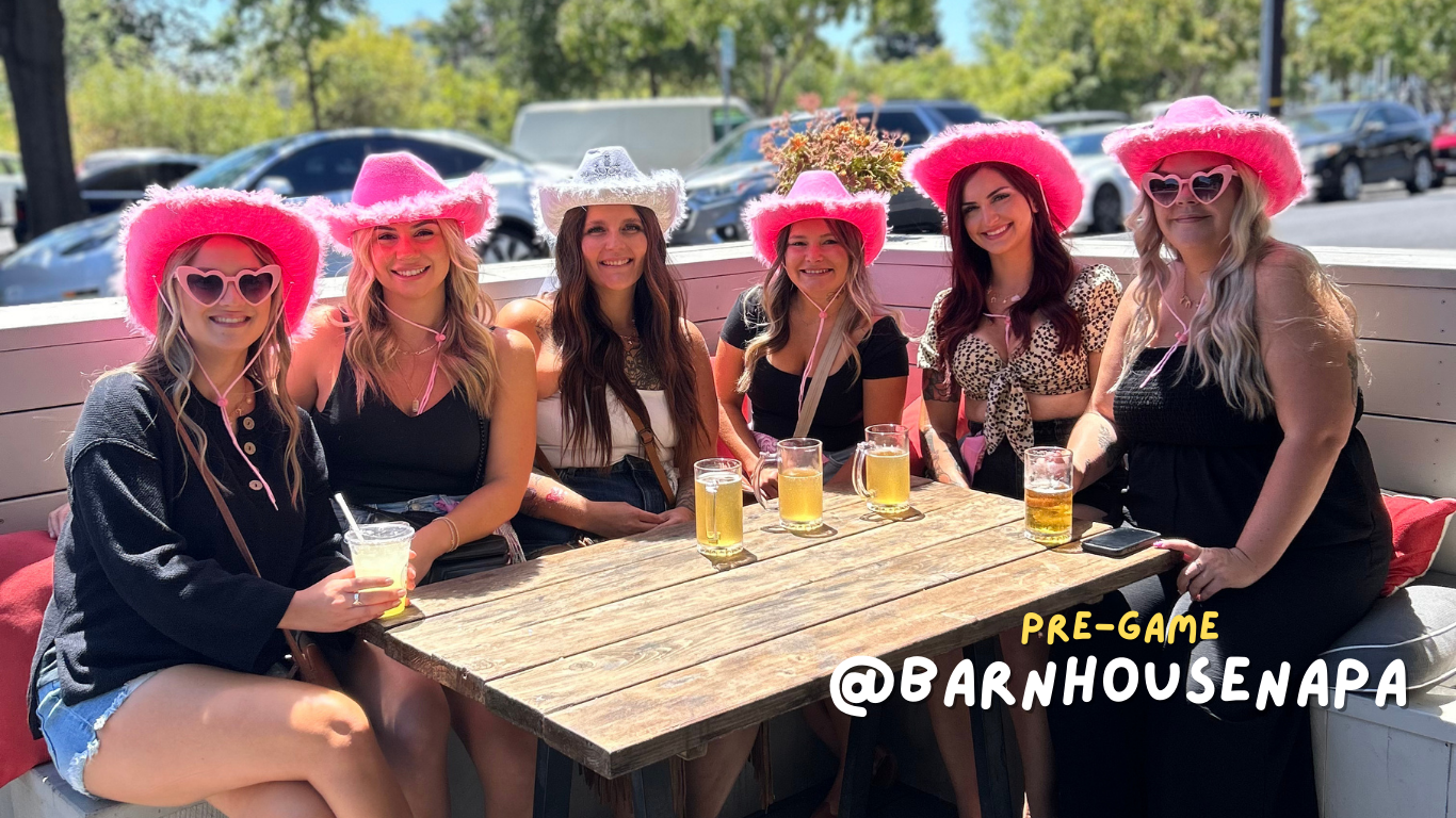 Six women wearing pink cowboy hats and sunglasses, sitting at a wooden table outdoors with drinks, smiling for the camera.