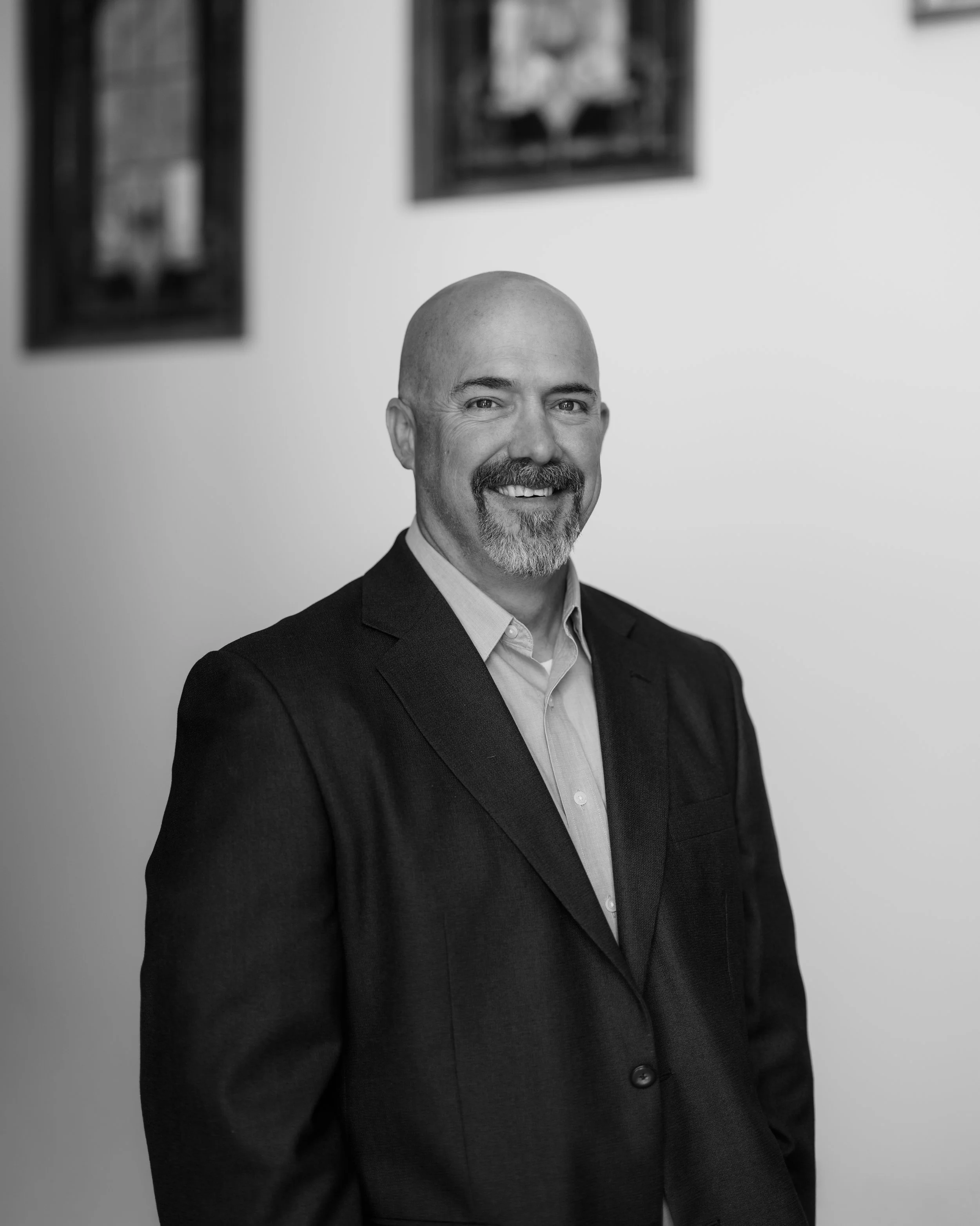 A middle-aged man with a beard and a shaved head, wearing a dark suit jacket and a checkered shirt, standing outdoors in a park with trees and a white building in the background on a clear day.