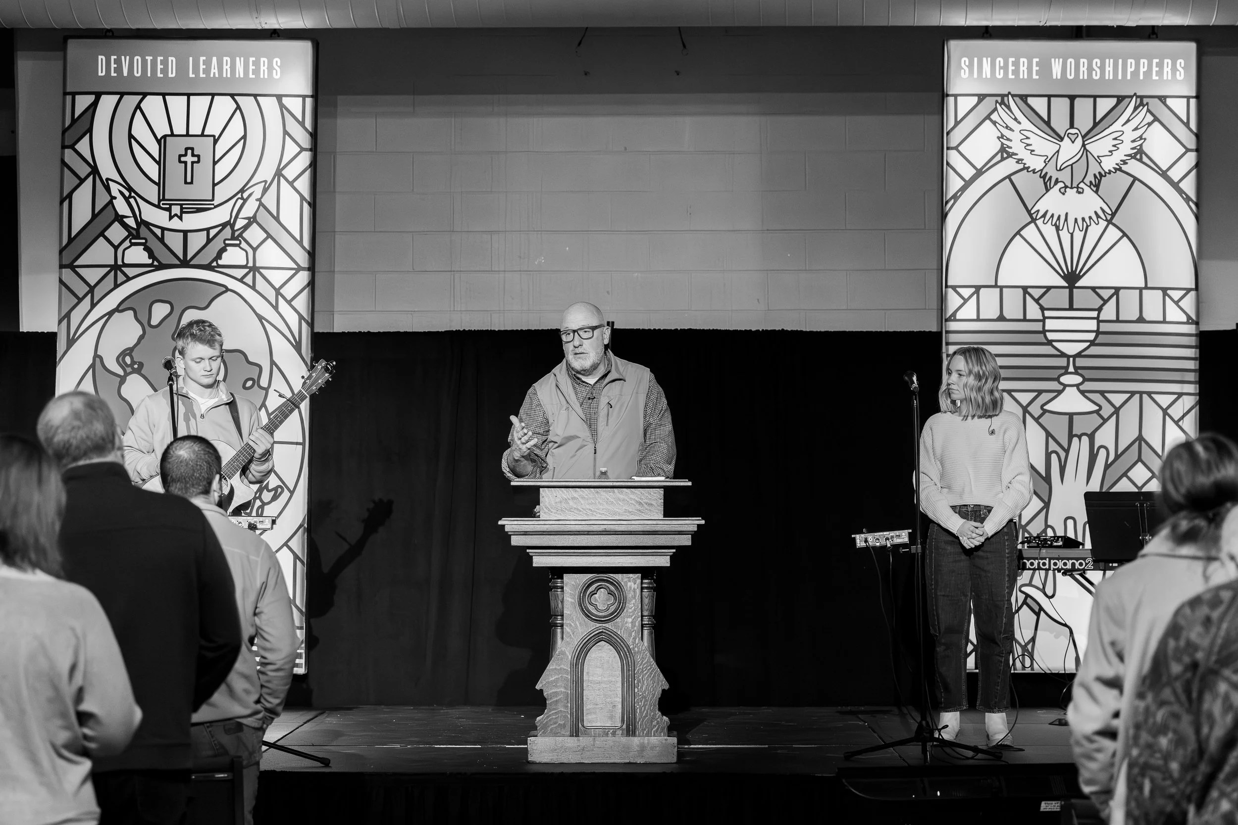 A church service with a preacher at the pulpit and two musicians on stage, one with a guitar and one with a keyboard, standing in front of religious banners that read 'Devoted Learners' and 'Sincere Worshippers'.