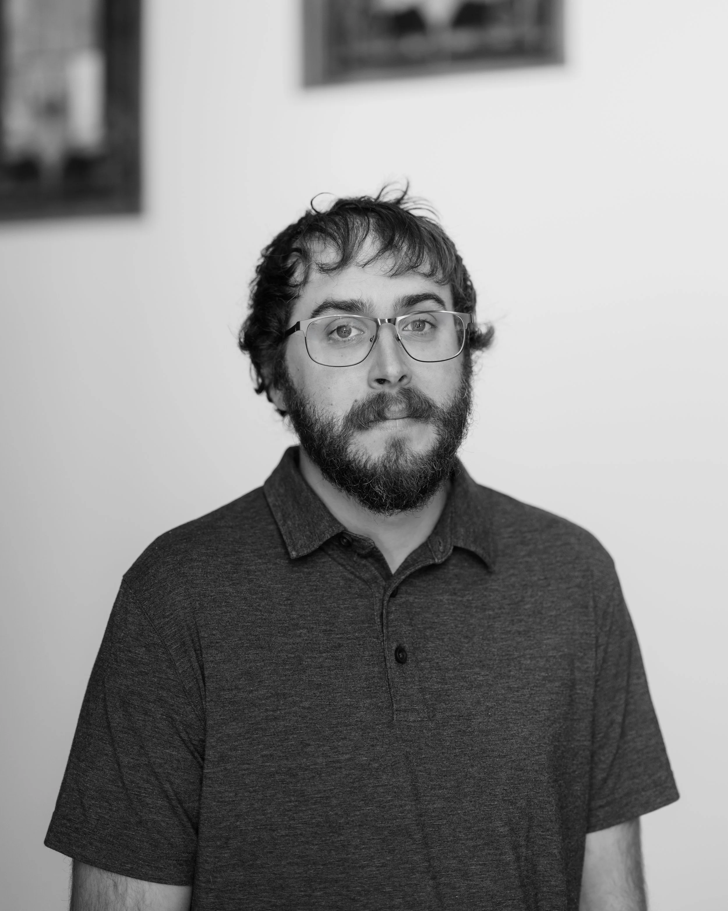 A man with glasses and a beard standing outdoors in front of a house with trees in the background.