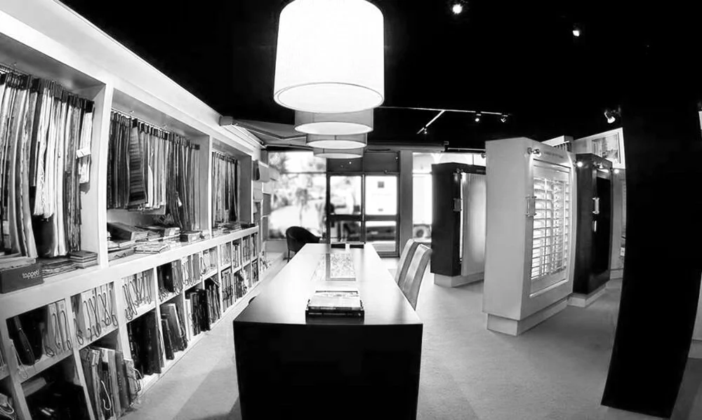 Interior of a clothing or fabric store with shelves of fabric samples on the left, a long table in the center, and display cases on the right.