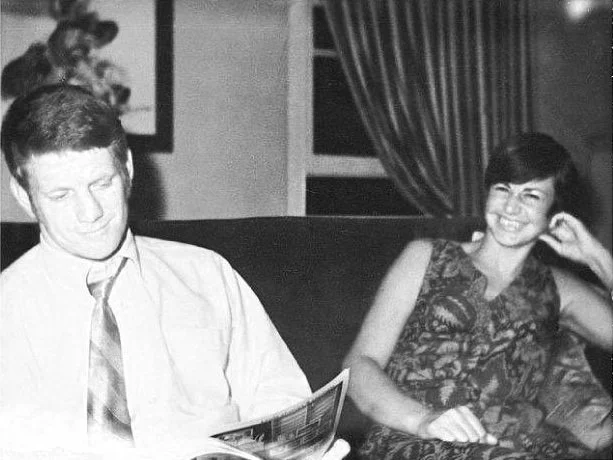 A black and white photo of a young man in a white shirt and tie reading a magazine, sitting next to a smiling woman in a patterned dress, sitting on a couch in a room with curtains.