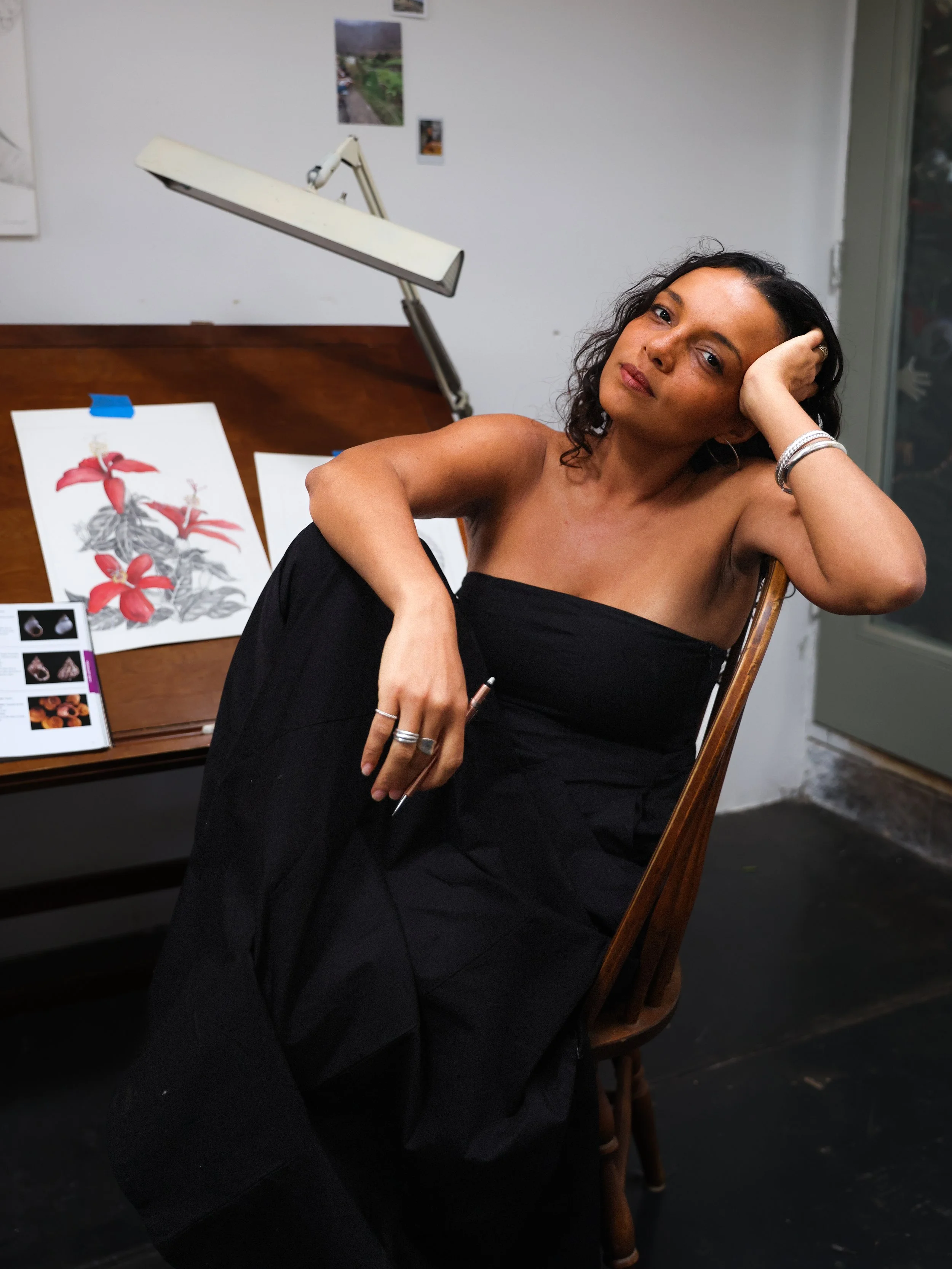 A woman with curly dark hair, dressed in black, sitting on a wooden chair, resting her head on her hand, in an art studio with paintings and photos on the wall, and a desk lamp overhead.