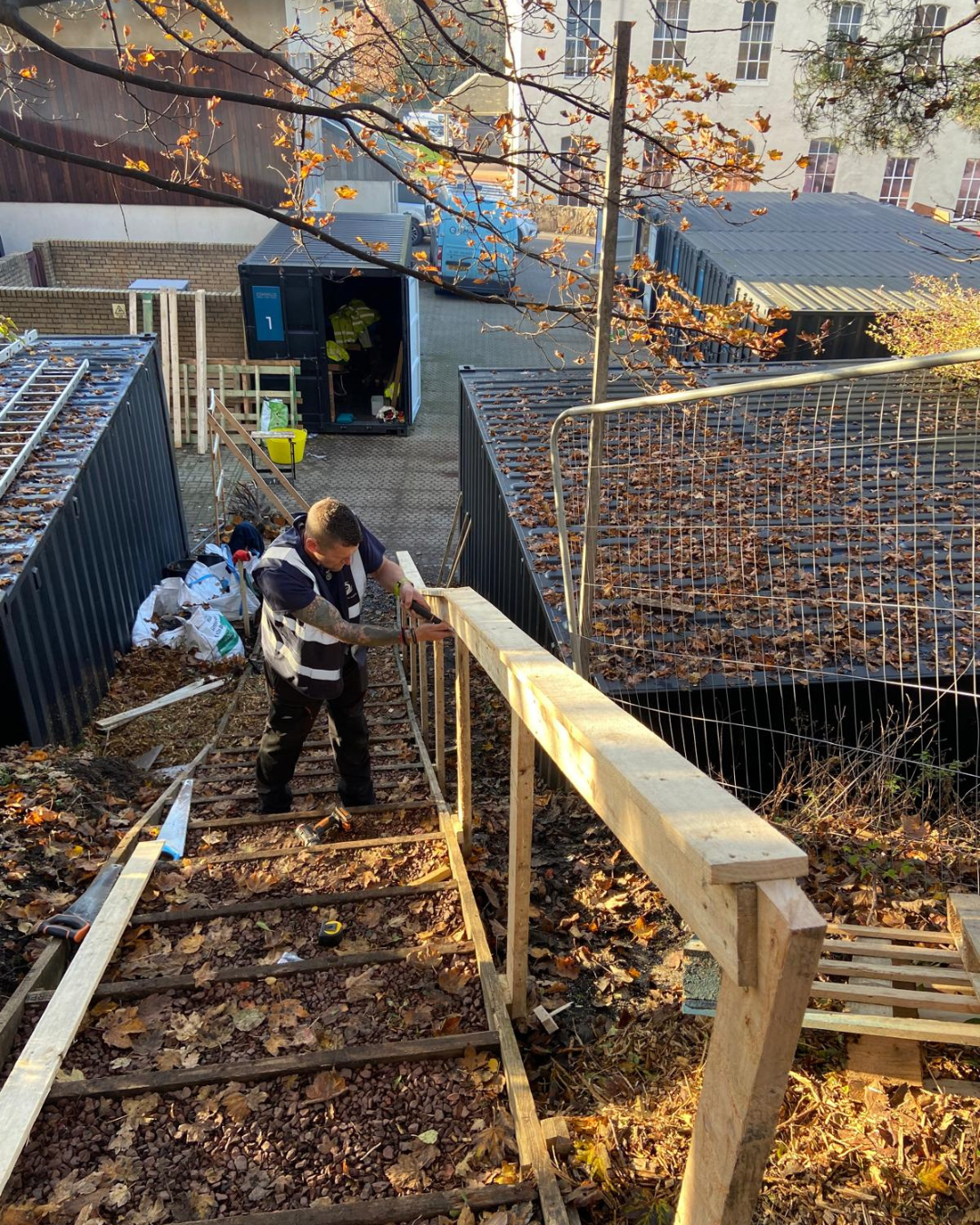A worker building a wooden railing on a staircase outdoors during autumn, with fallen leaves on the ground and a construction trailer in the background.