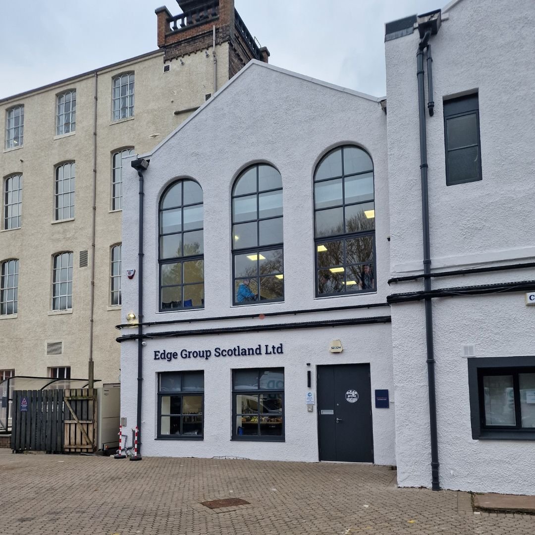 The white building has the sign 'Edge Group Scotland Ltd' on it, with large arched windows on the upper floor and smaller rectangular windows on the ground floor, situated on a paved area.