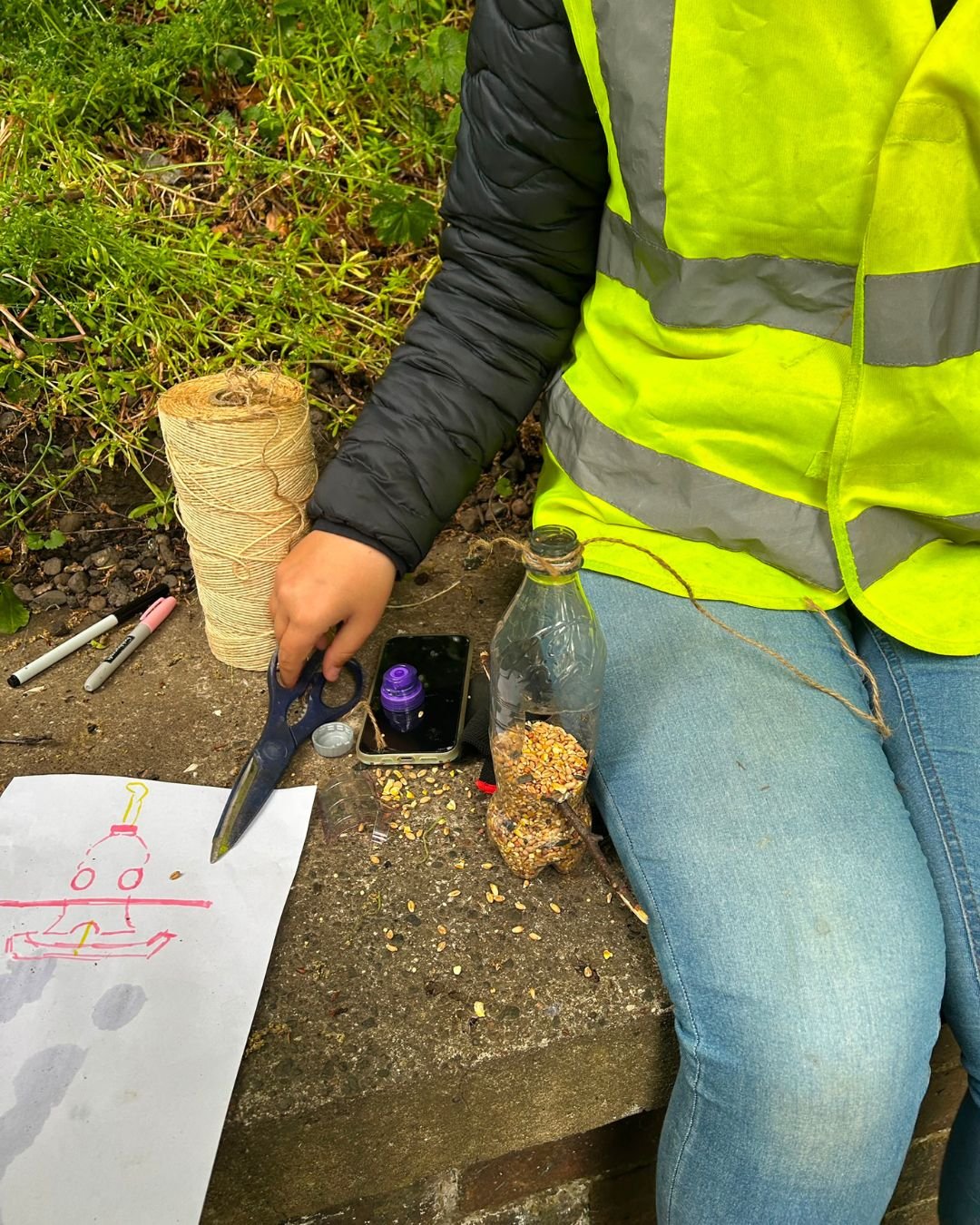 Person wearing a high-visibility safety vest and jeans sitting on a concrete surface, engaged in an outdoor activity involving making bird feeders, and a drawing of a bird feeder on paper, surrounded by markers and other materials.