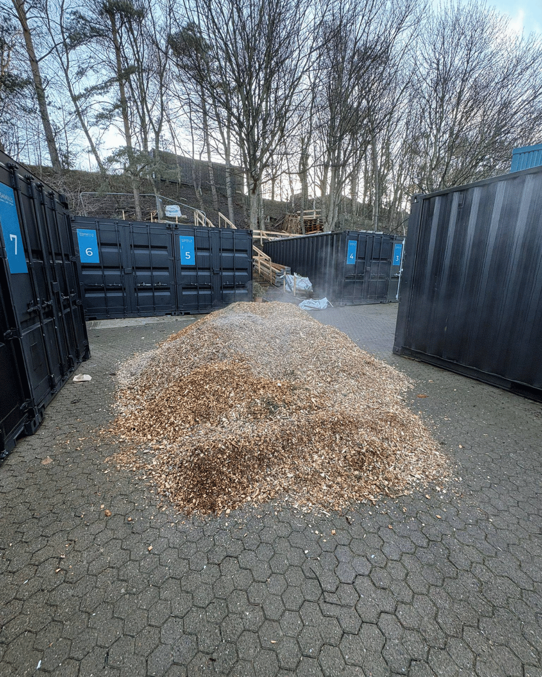 A large pile of wood chips or mulch in an outdoor area with black containers in the background. There are trees and a hillside with trees in the background.