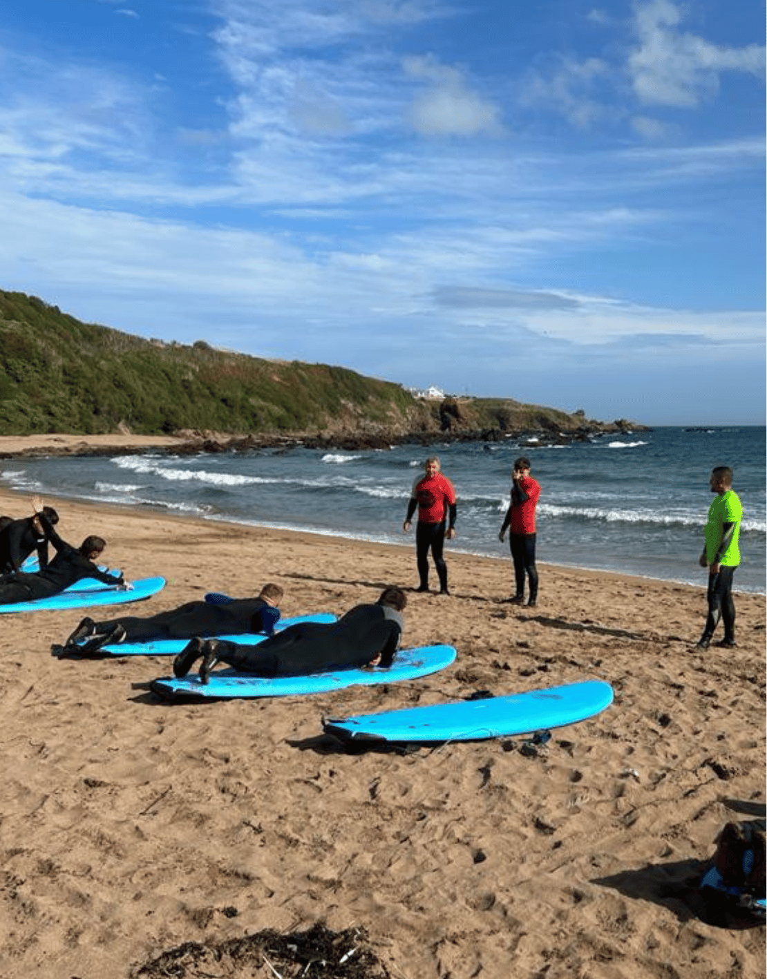 People lying on blue surfboards on a sandy beach in front of instructors, with others standing and talking near the water, ocean waves in the background, and a partly cloudy sky.
