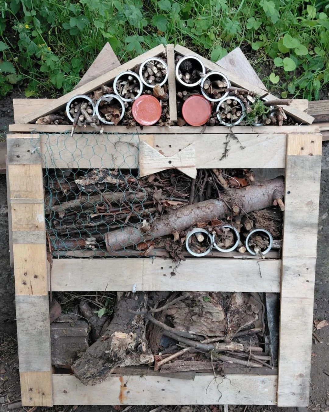 A wood insect hotel made from a wooden pallet with various compartments filled with natural materials like sticks, bark, logs, and twigs, some metal rings and clay pots, and a wire mesh section, set outdoors surrounded by green foliage.