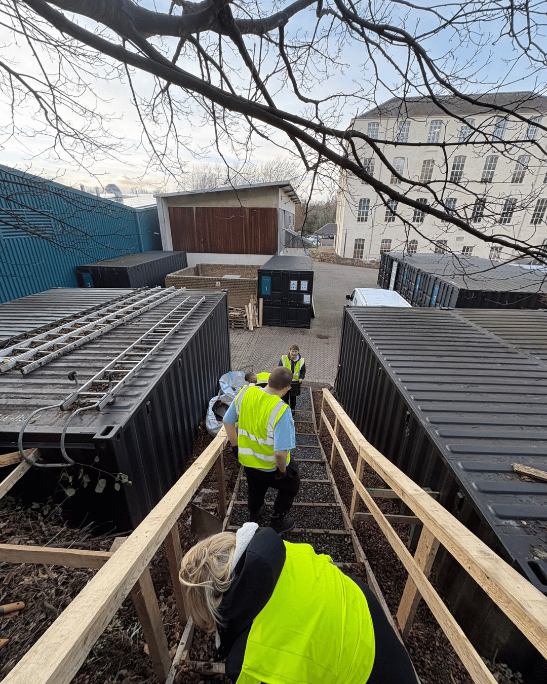 Workers in high-visibility vests walking down a set of stairs at a construction site, with large shipping containers and a brick building in the background.