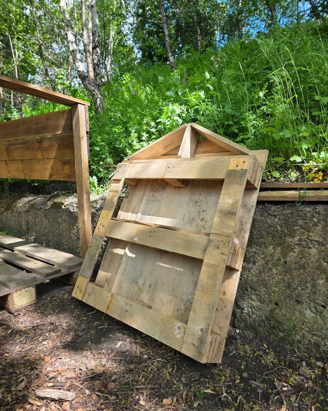 Unfinished wooden garden planter frame leaning against a rock wall outdoors, with greenery and trees in the background.