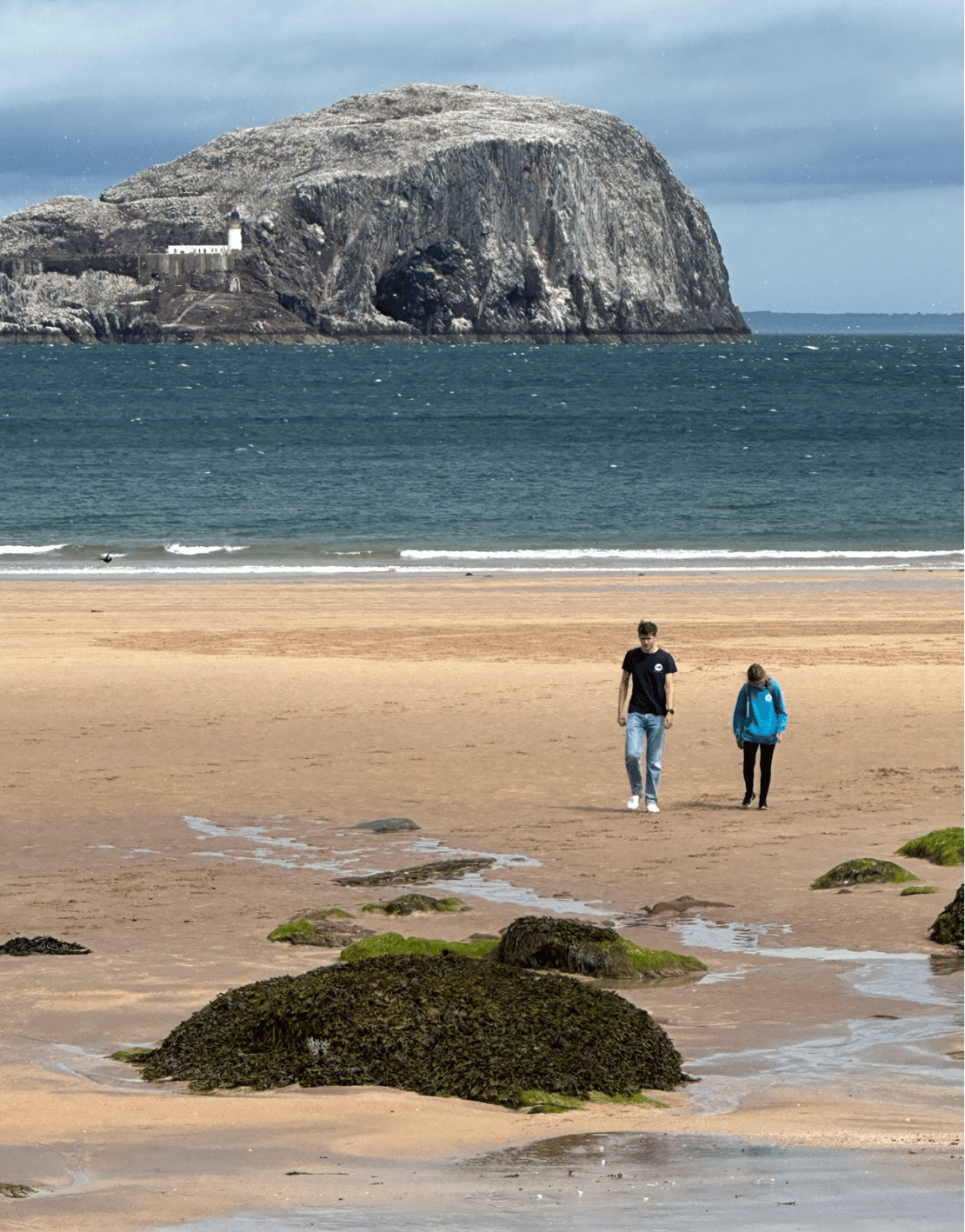 Two people walking on a sandy beach near the water, with a large rocky cliff or island in the background and moss-covered rocks in the foreground.