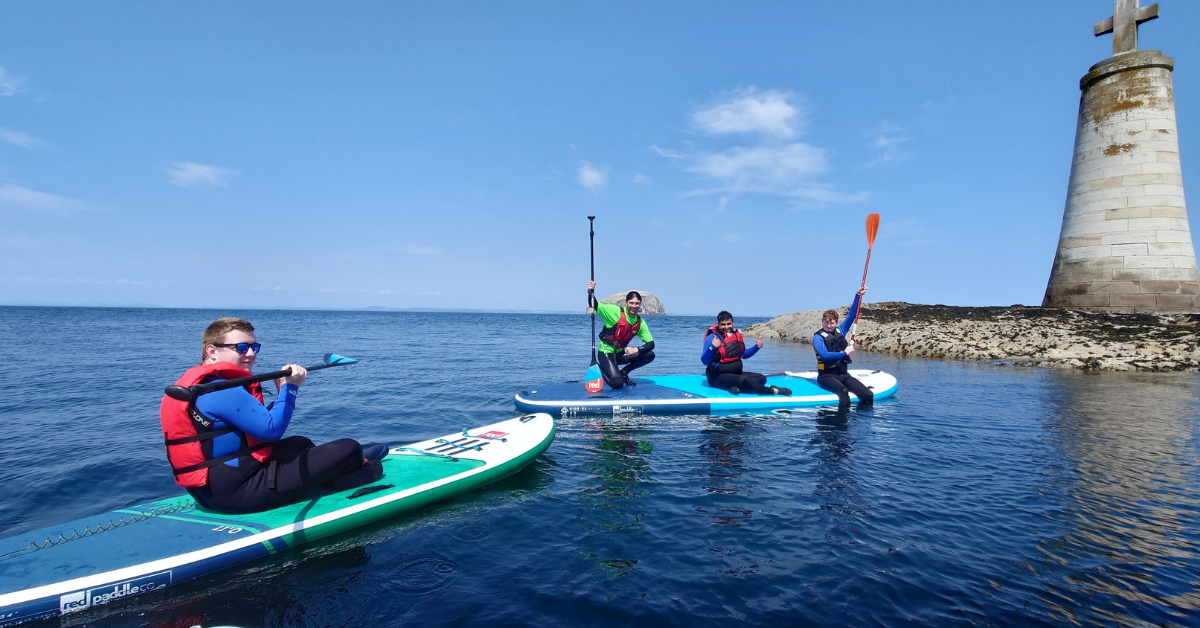 Four people paddleboarding in the ocean near a rocky shore with a lighthouse.