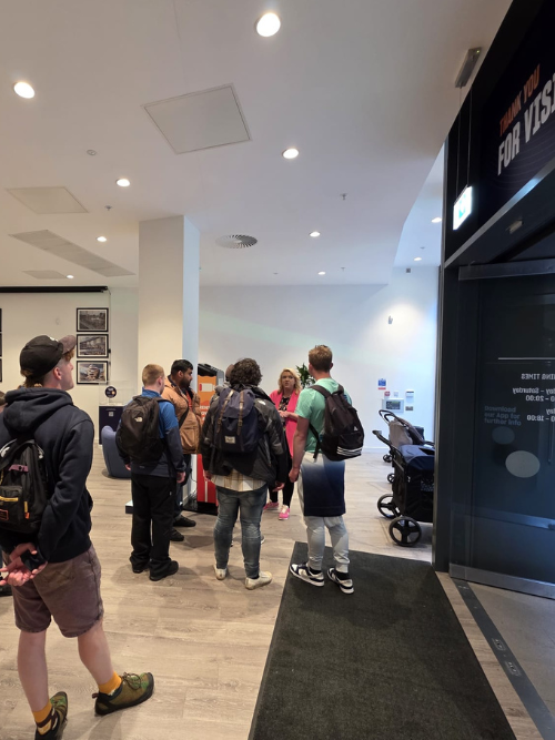 Group of young people, mostly males, standing in line at an airport security checkpoint, with a TSA agent and a stroller nearby.