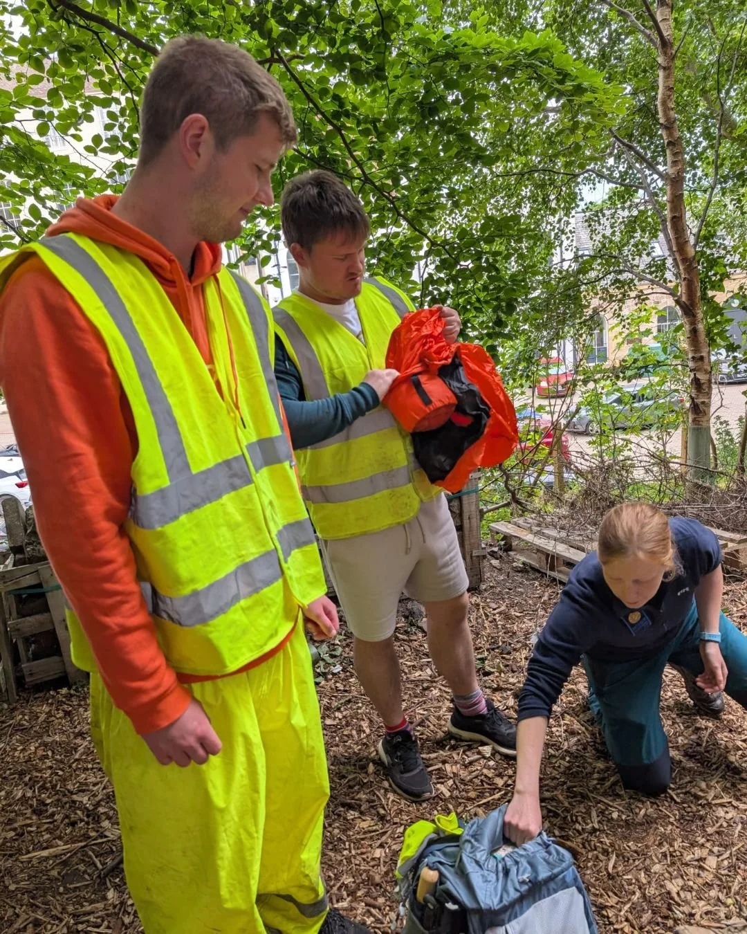 Two men and one woman in a wooded area, with the women reaching into a backpack on the ground and the men looking on, all wearing high visibility safety vests.
