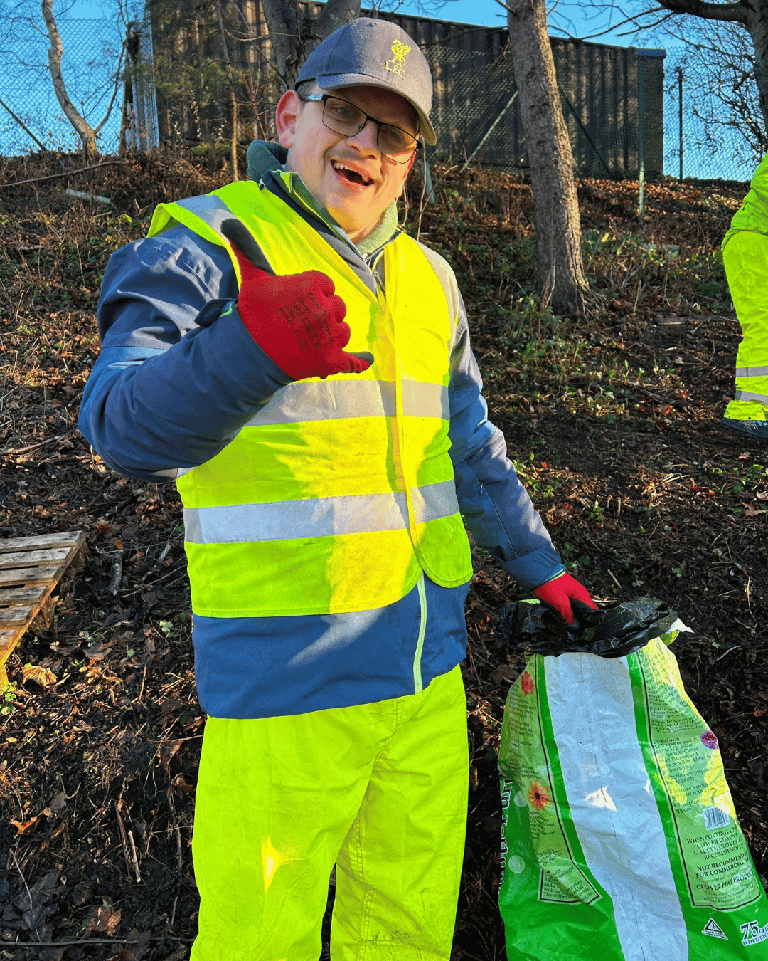 Man wearing a yellow safety vest, gray and blue jacket, red gloves, and a gray cap with a yellow emblem, standing outdoors on a dirt and leaf-covered hill with trees and a black fence in the background. He is smiling and making a shaka sign with his 