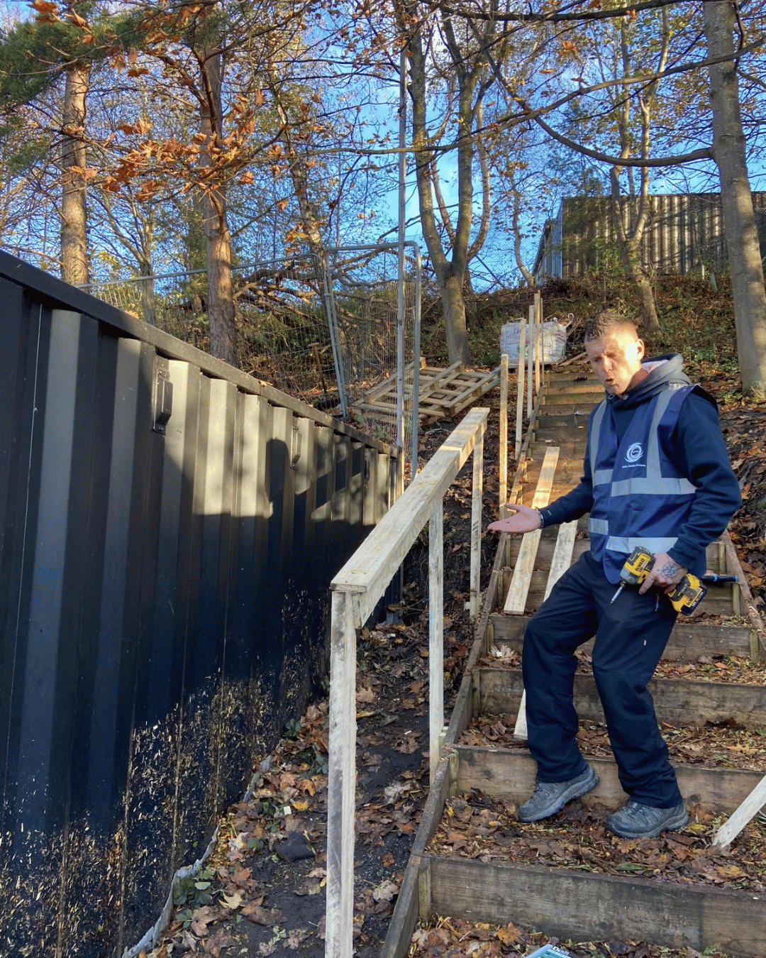 A man wearing a blue safety vest and holding a drill stands on a staircase under construction, explaining the ongoing work on a wooden handrail, next to a black metal container on a hillside surrounded by trees with autumn leaves.