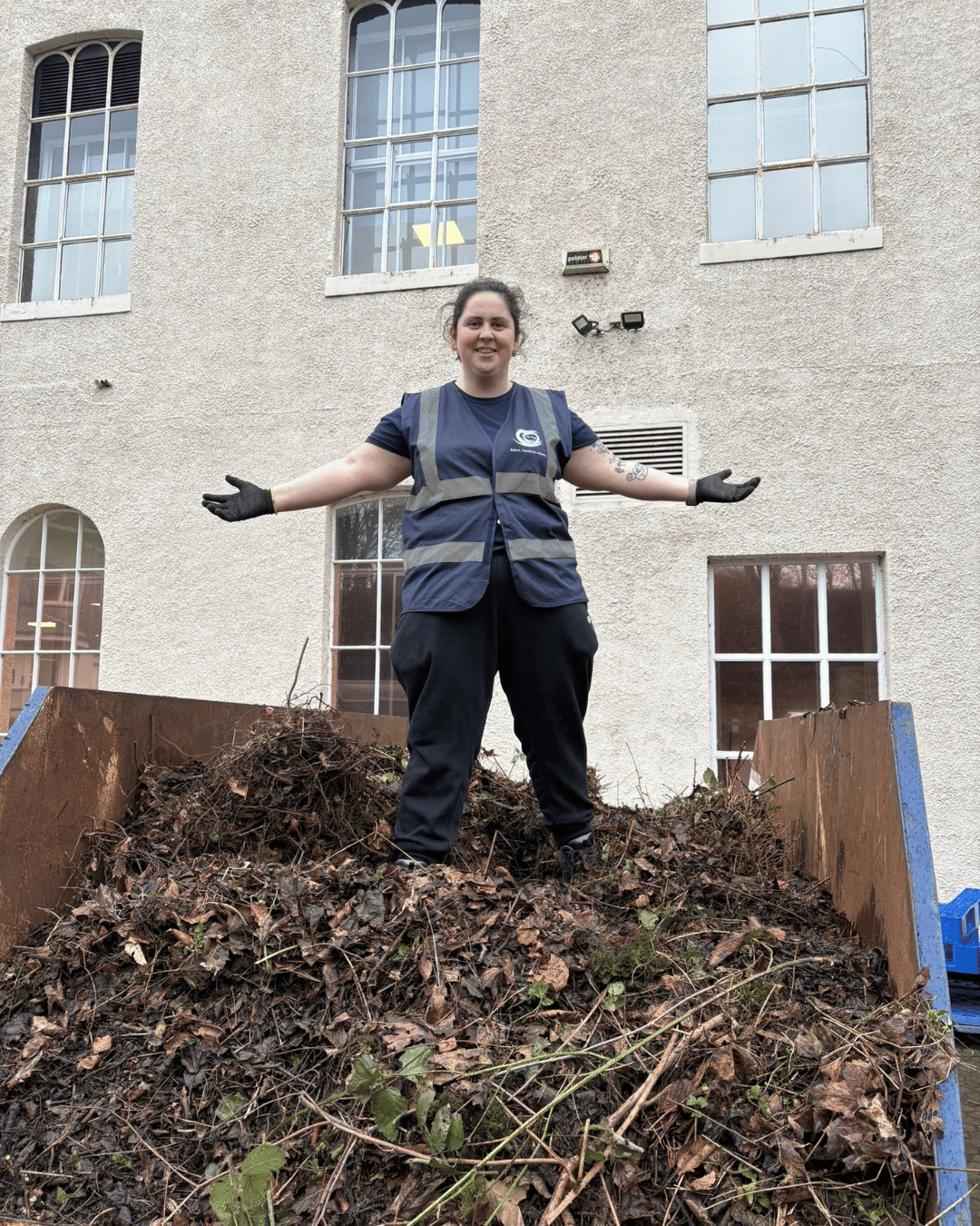 A woman standing on a pile of dried leaves and branches, with arms outstretched, in front of a beige building with large windows.