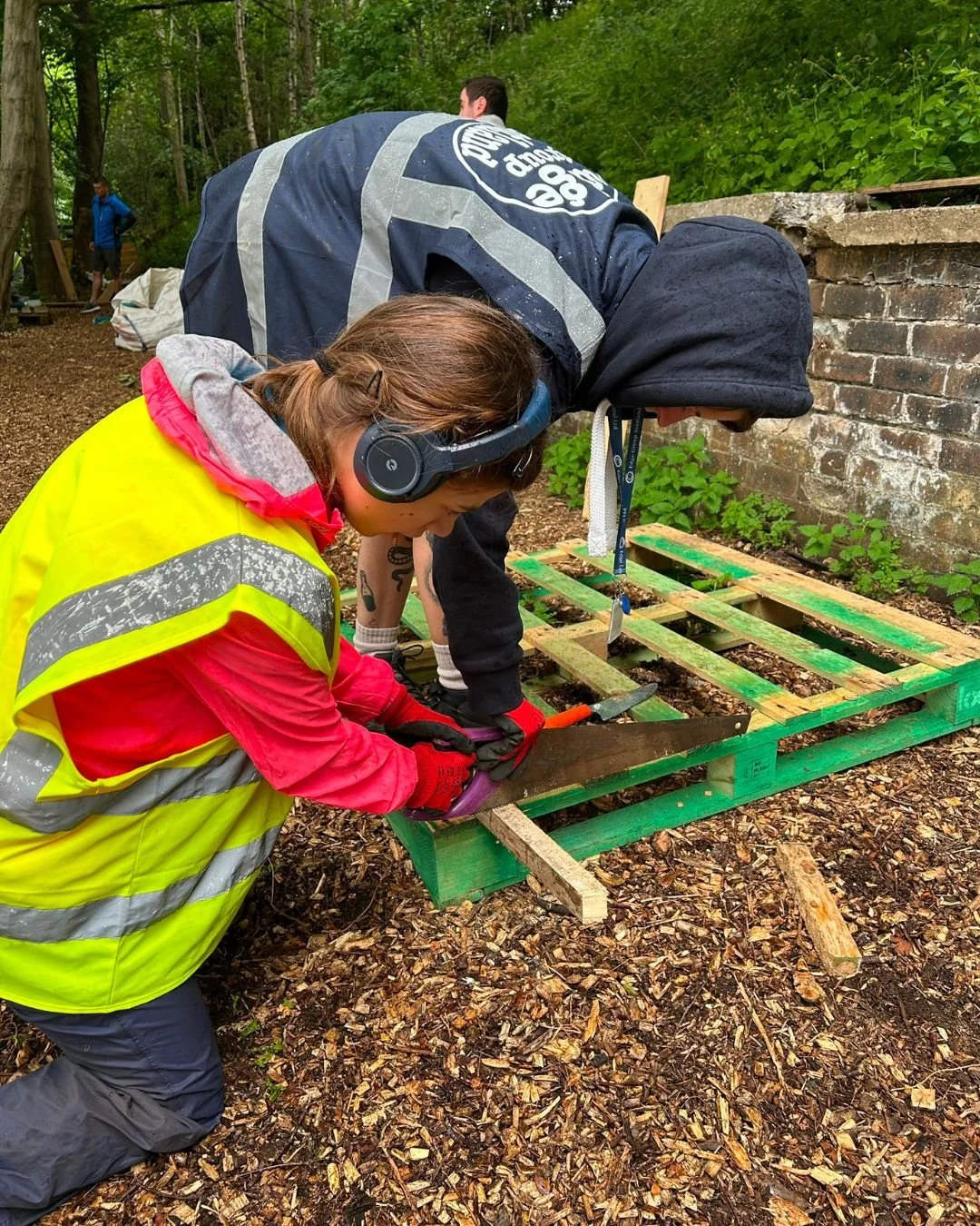 Two people working together outdoors on a woodworking project in a forested area, with one person wearing a yellow safety vest and the other in a dark hoodie, using a saw on a green wooden pallet.