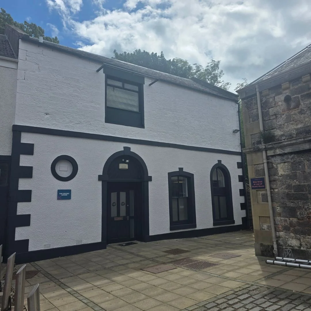White building with black trim, arched windows, and a black door, situated on a paved street with a stone wall nearby, under a partly cloudy sky.