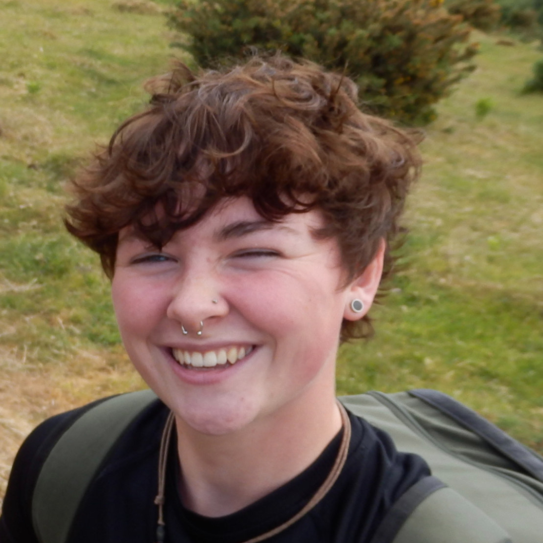 A smiling young person with curly brown hair, wearing a nose ring, earrings, a black shirt, and a backpack, outdoors with green grass and bushes in the background.