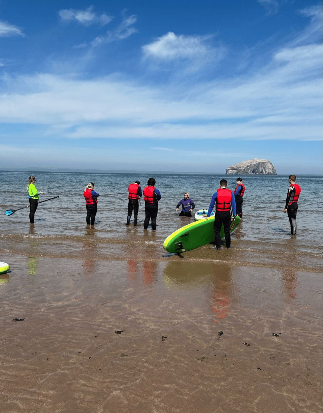 Group of people in life jackets on the beach preparing for a paddleboarding lesson, with a small island in the distance under a partly cloudy sky.