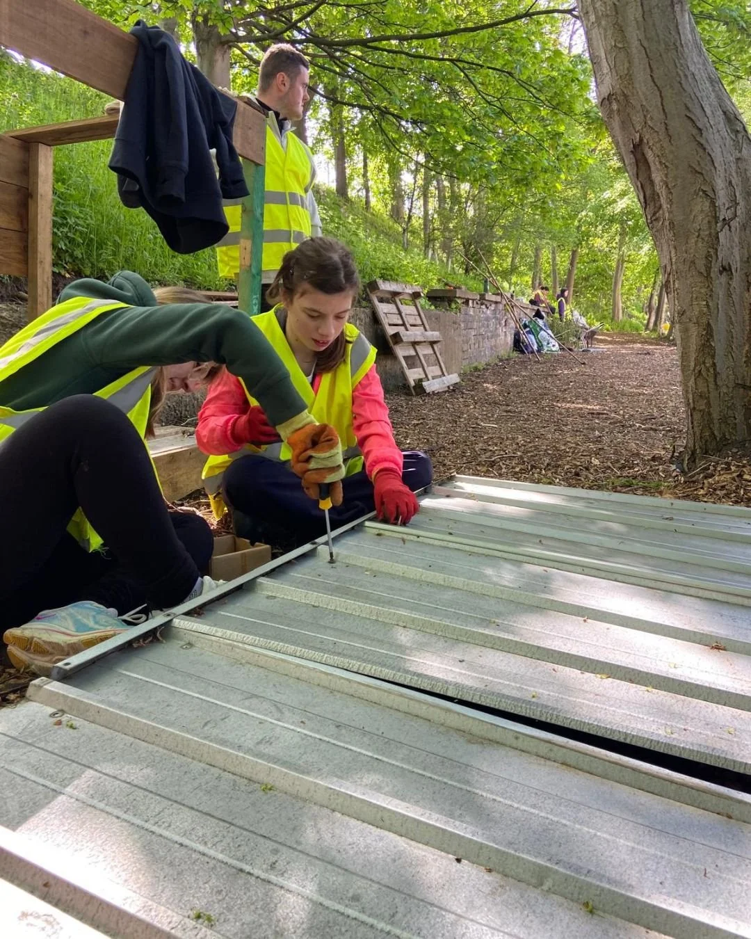 People working outdoors on a metal roof, wearing yellow safety vests, one person screwing a panel, others assisting, surrounded by trees and greenery.