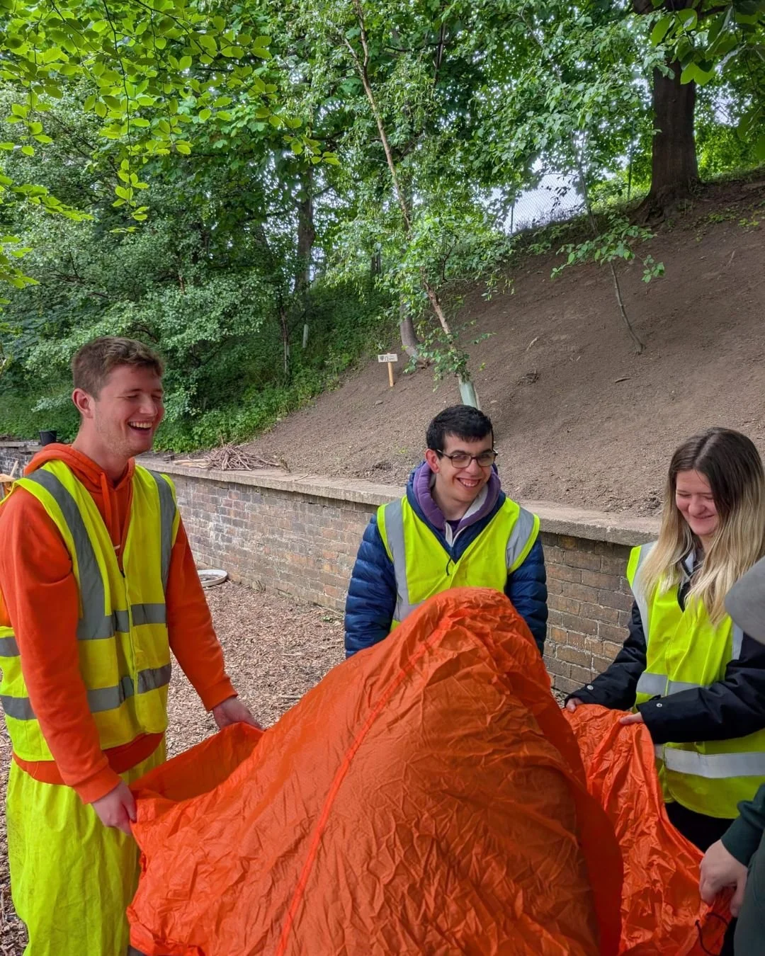 Four young people wearing yellow safety vests holding an orange tent outdoors, smiling and laughing. They are standing on a dirt path along a brick wall with a wooded hillside in the background.