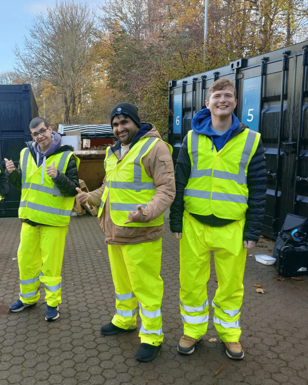 Three young men wearing high-visibility yellow safety vests and pants, standing outside on a paved area, smiling and giving thumbs-up signs, with trees and black storage containers in the background.