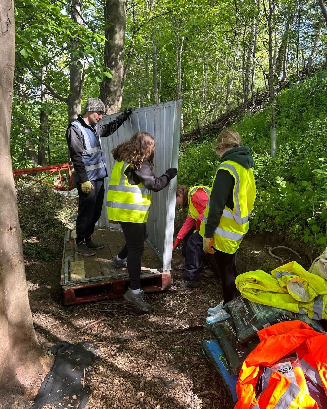 Group of people wearing yellow safety vests working together to install or repair a metal fence in a wooded area during daylight.