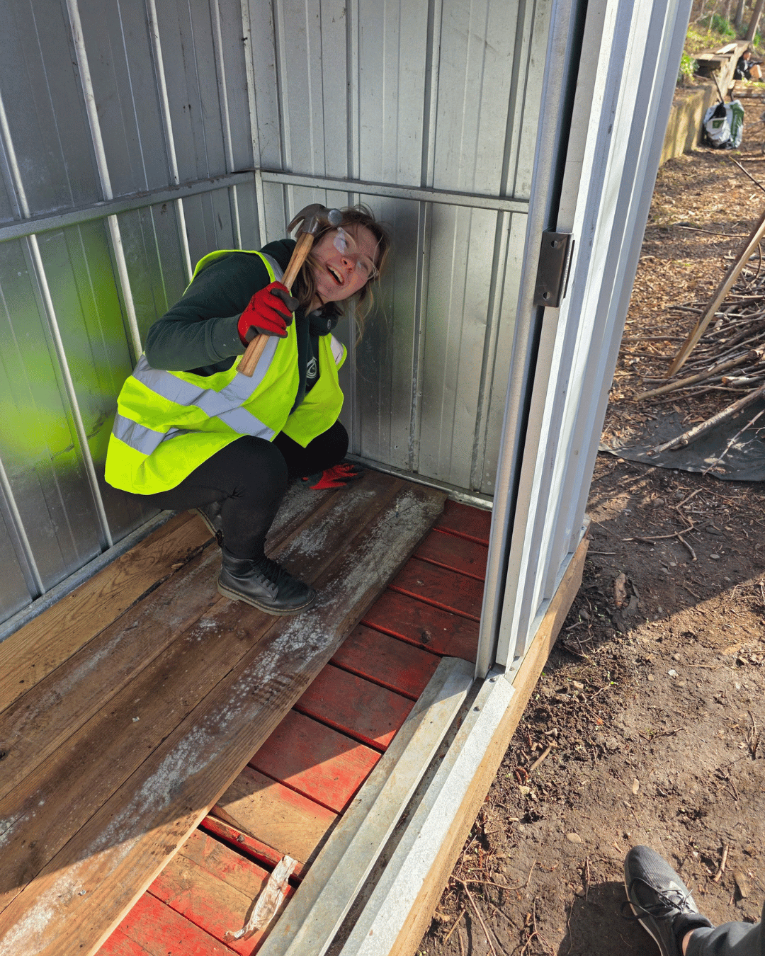 A woman smiling inside a small metal structure on a wooden platform, holding a hammer, wearing safety glasses, a high-visibility vest, black gloves, and black outdoor boots, standing on a construction site.