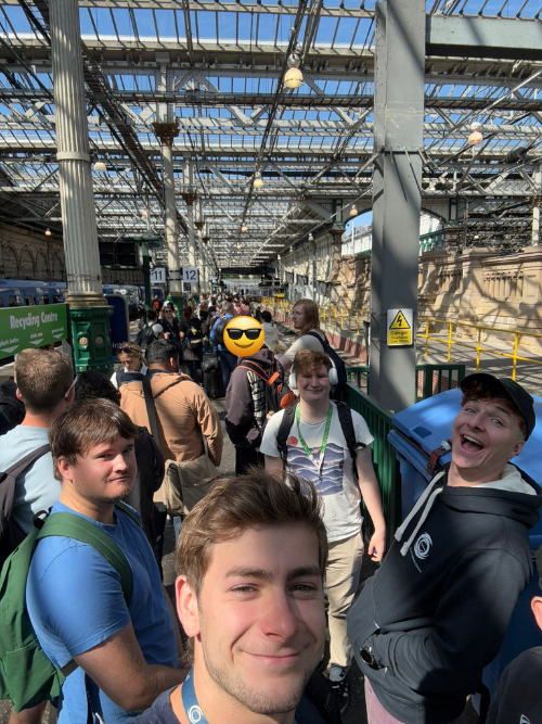 Group of young people waiting in line at a train station with glass ceiling, metal beams, and people standing with backpacks.