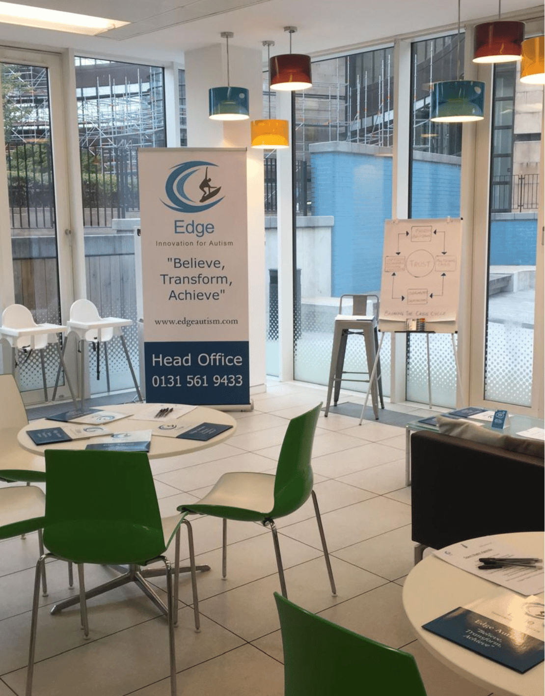 Interior of a meeting room with tables, chairs, a banner with the Edge Autism logo, and a whiteboard with diagrams.