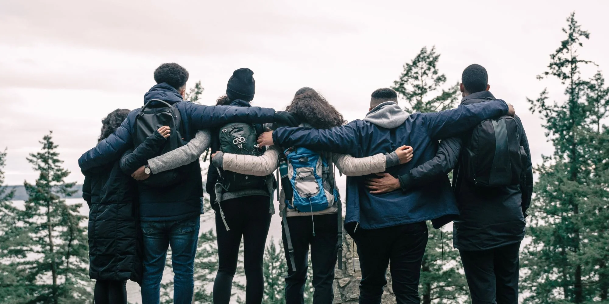 Six hikers with backpacks standing arm-in-arm on a mountain top, overlooking a forested landscape.
