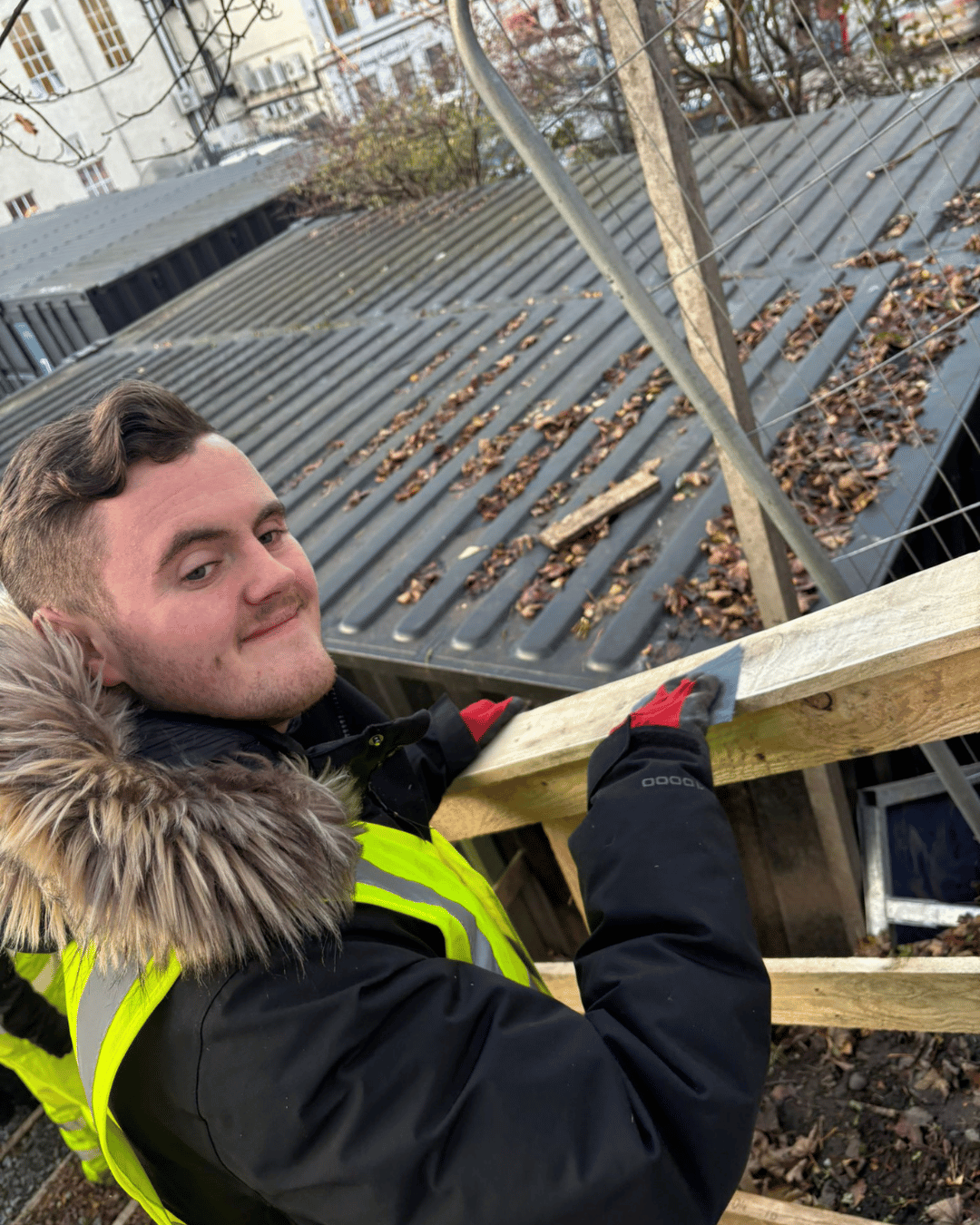 A young man wearing a yellow safety vest and gloves is installing wooden beams on a construction site outdoors, with a sloped metal roof and leafless trees in the background.