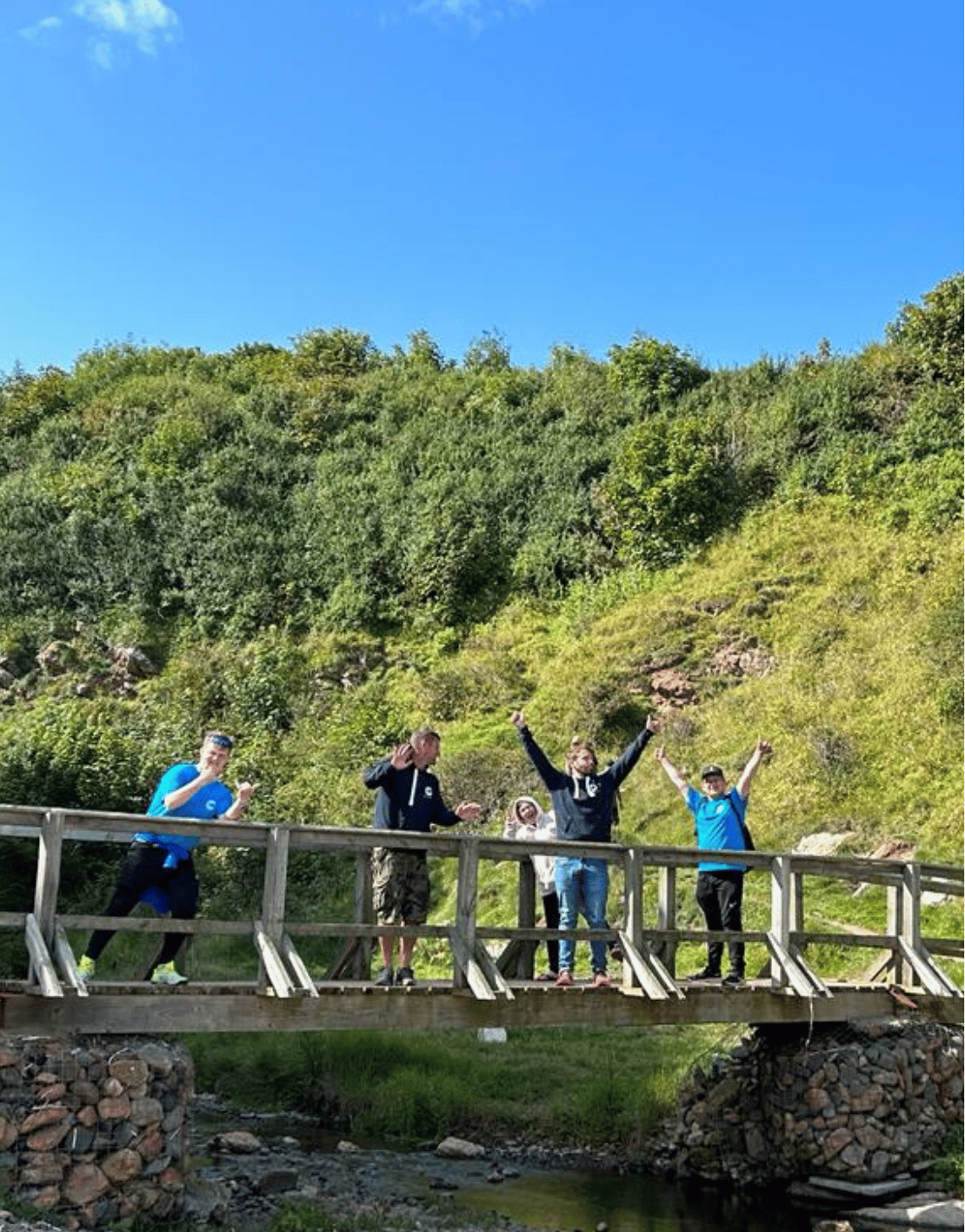 Five people standing on a wooden bridge, waving and smiling, with a green hillside and clear blue sky in the background.