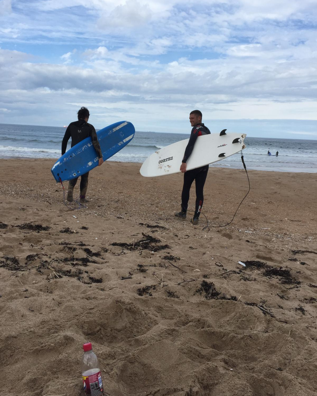 Two surfers walking on the beach holding surfboards, one with a blue board and the other with a white board. The sky is partly cloudy and the ocean waves are in the background, with a few people surfing in the distance. There is a bottle in the sand in the foreground.