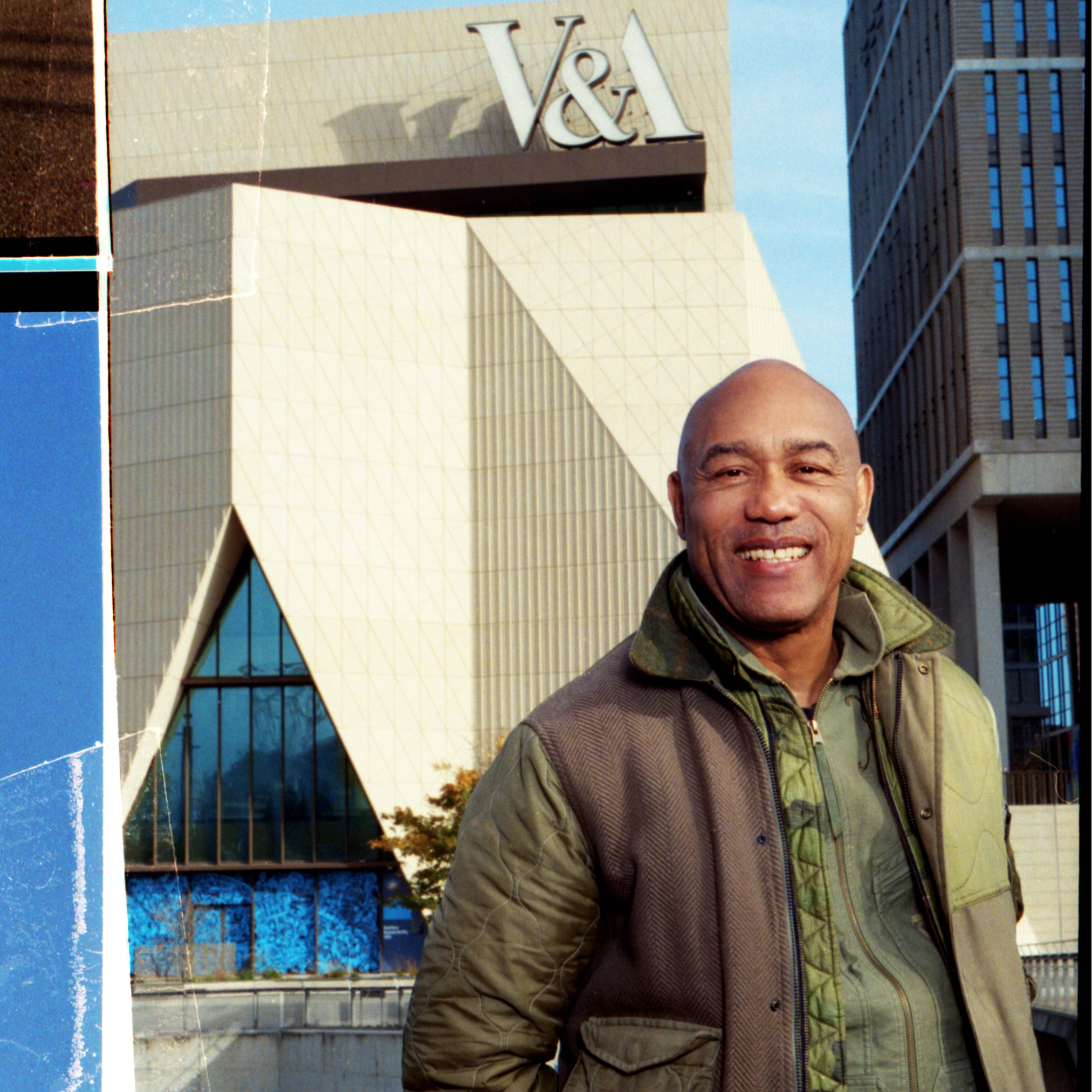 Person standing outdoors in front of the V&A Dundee building, with the museum’s angular, modern architecture visible behind them. The building features distinctive geometric forms, textured concrete surfaces and large glass sections.
