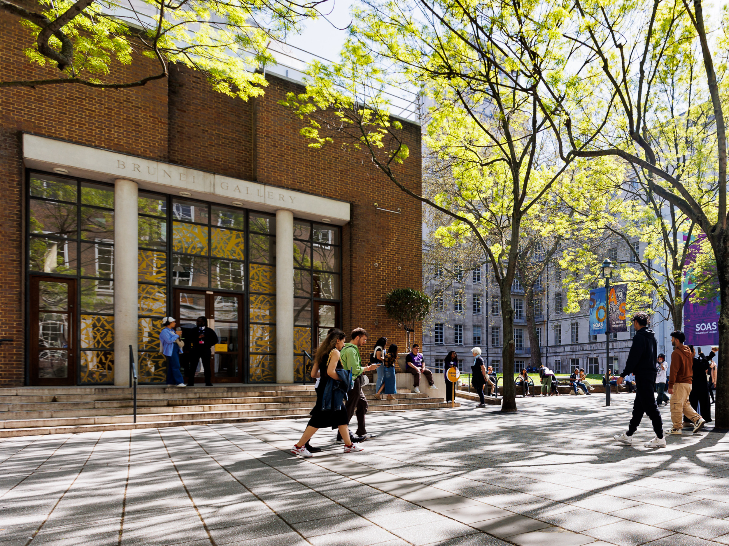 People walking and gathering outside the Brunei Gallery at SOAS, with a brick building, large glass entrance, spring foliage on trees, and campus banners visible in a sunlit courtyard.