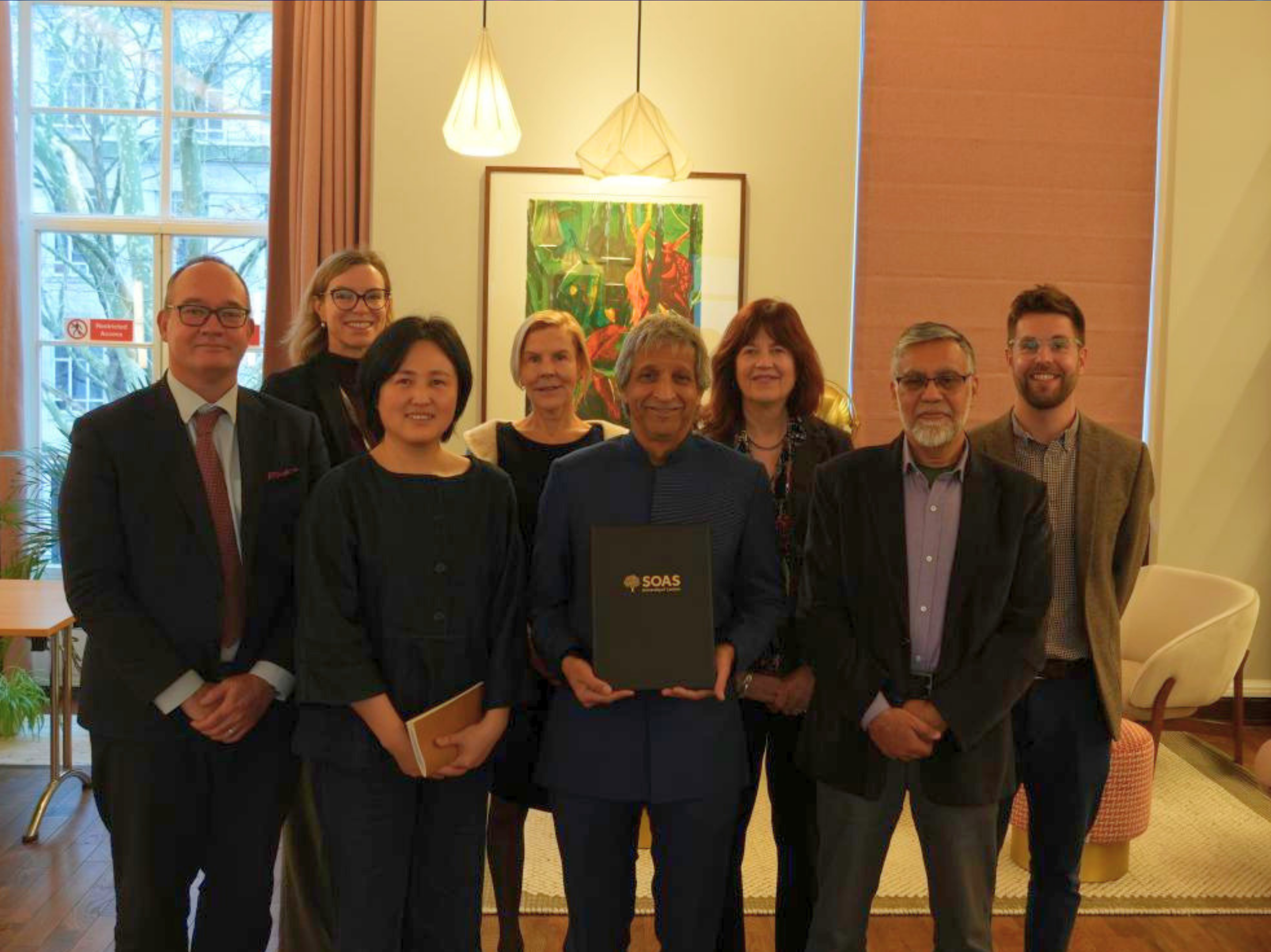 Group of people standing together indoors in front of a framed artwork, with the central person holding a folder displaying the SOAS logo, in a softly lit room with pendant lights and large windows.
